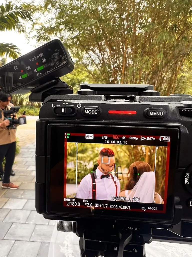 Sony cinema camera on tripod recording a bride and groom outdoors; LCD shows 4K 59.94p, AF on the groom, wireless audio on the rig