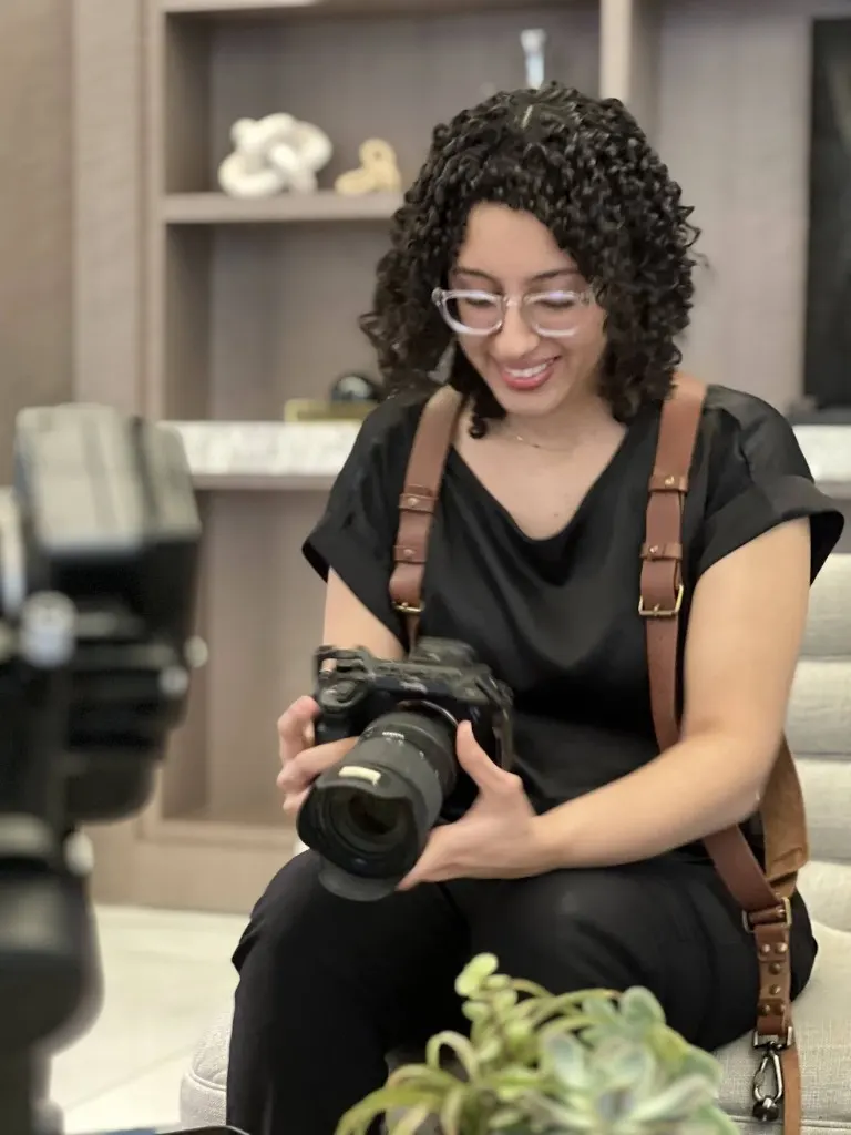 Maria Peña reviewing shots on a professional camera indoors, wearing a dual-camera leather harness with a second rig visible in the foreground